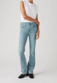 Jeans acampanados de color azul claro con una cintura de altura media, hechos de denim. Combinados con un top sin mangas blanco, modelo que lleva zapatos negros de punta cuadrada.