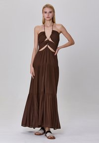 Robe maxi marron avec des détails découpés et un col halter, présentant un design fluide à volants et une texture douce ; associée à des sandales simples.