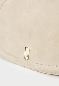 Bolsa de camurça bege com forma curva, apresenta um detalhe retangular em tom dourado e costura branca contrastante ao longo da borda.