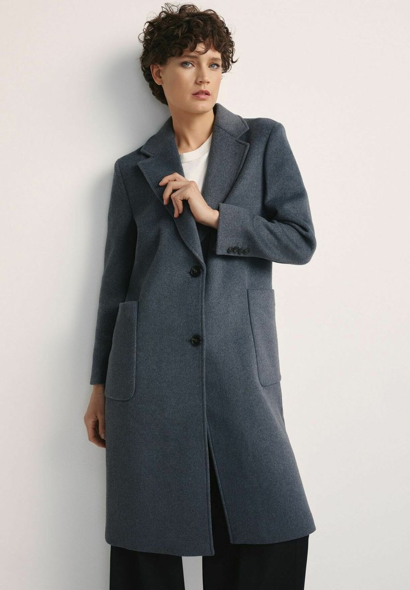 Kobieta o krótkich, kręconych włosach, ubrana w długi, szary płaszcz, pod który ma na sobie białą koszulę i czarne spodnie, stoi na tle jednolitego białego tła.