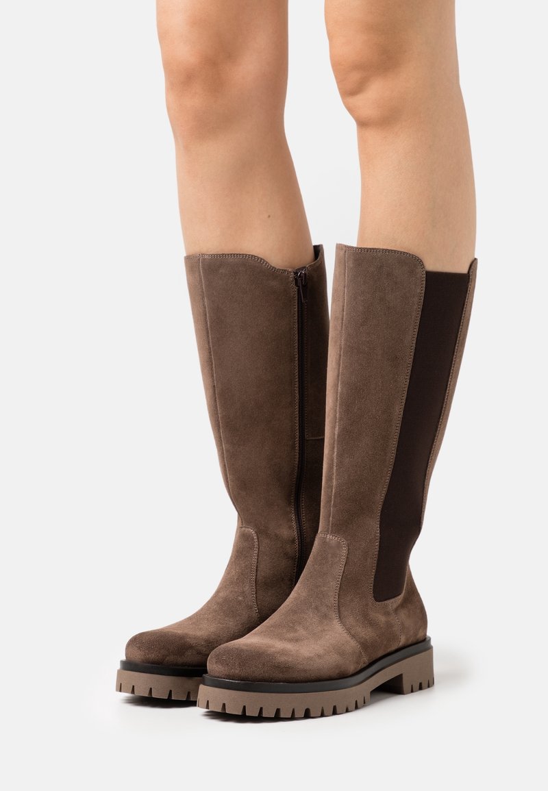 Bottes en daim marron montantes jusqu'au genou avec semelles épaisses et panneaux élastiques latéraux, portées sur des jambes nues sur un fond uni.