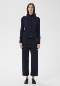 Tengerészkék hosszú ujjú kardigán, magas nyakkal és gombos lezárással, sötétkék egyenes szárú farmerral és fekete fűzős cipővel párosítva.