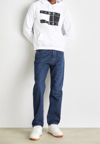 White hooded sweatshirt with black "TOMMY JEANS" graphic, paired with blue denim jeans and white sneakers with tan soles.