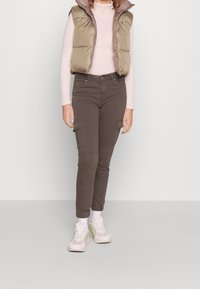 Olivový puffe vest přes světle růžový proužkovaný top, zkombinovaný s tmavě hnědými cargo kalhotami s bočními kapsami a béžovými teniskami se zelenými akcenty.