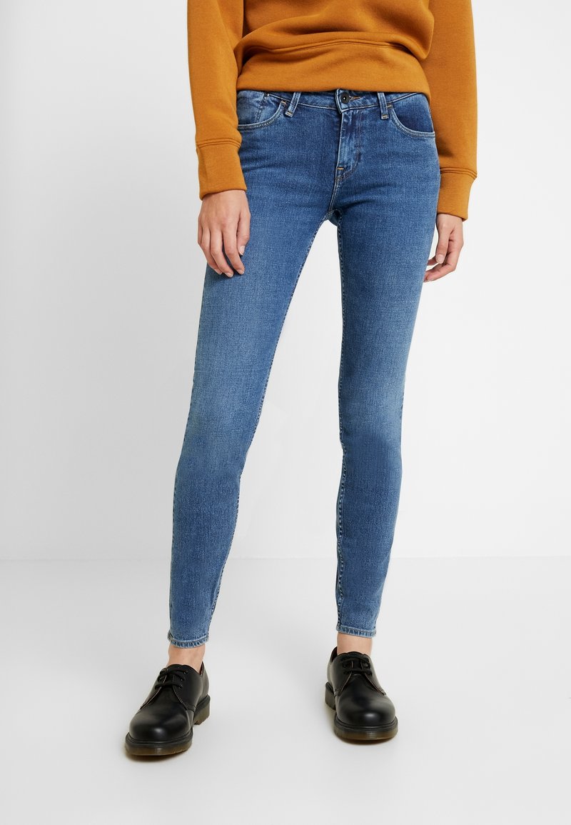 Skinny blue jeans in denim fabric, featuring a mid-rise waist, five pockets, and a classic button fly. Paired with black lace-up shoes.