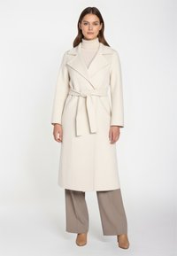 Cappotto lungo color crema con vita cintrata e rever a scollo a V. Presenta maniche lunghe e una silhouette dritta, abbinato a pantaloni color taupe.