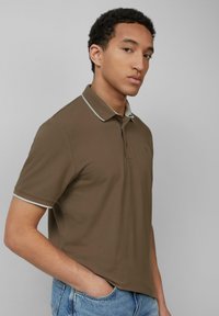 Braunes Poloshirt mit kurzem Kragen, kontrastierenden hellen Abschlüssen und einem gestickten Logo auf der linken Brust. Getragen mit hellblauen Jeans.