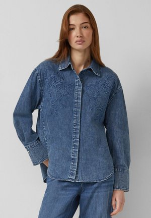 Kobieta o rudych włosach, ubrana w niebieską dżinsową koszulę i dżinsy z delikatnym kwiatowym haftem na piersi, stoi na jednolitym szarym tle.