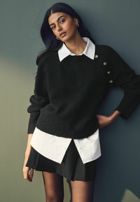 Sort strikket sweater med ribbet kant, hvid skjorte lagt under, og en plisseret sort nederdel. Guld detaljer med knapper.