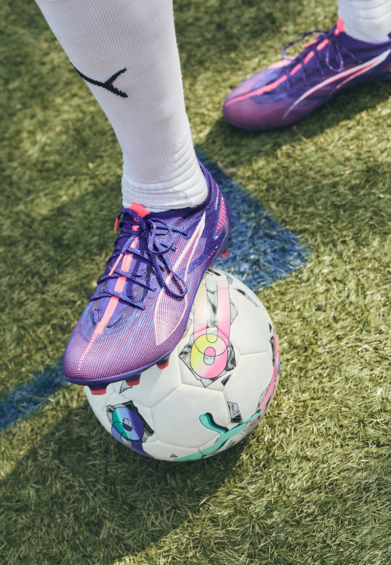 Lila fotbollssko med strukturerad yta och rosa accenter vilar på en färgglad, mönstrad fotboll, på grön konstgräs.