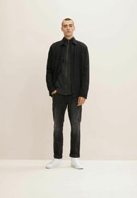 Model v černé košili na knoflíky přes šedý svetr na zip, doplněný tmavými džíny a bílými teniskami, na neutrálním pozadí.