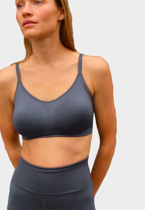 Kvinde iført en grå sports-bh og højtaljede leggings, der kigger let opad med hænderne bag ryggen mod en ensfarvet baggrund.