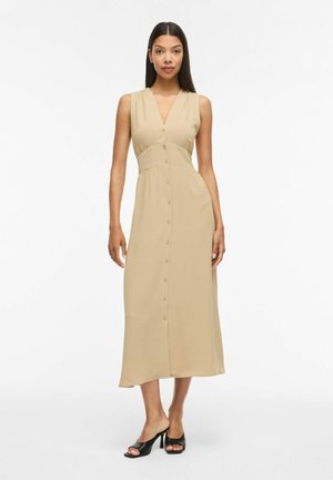 Femme portant une robe midi beige sans manches à boutons sur le devant et col en V, associée à des mules noires à talons ouverts sur fond blanc.