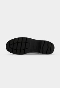 Semelle de chaussure noire avec motif de bande de roulement rainuré et indentation rectangulaire au milieu sur un fond clair uni.
