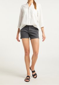 Valkoinen pitkähihaisellinen paita ilman nappeja kauluksessa, yhdistettynä tummanharmaisiin shortseihin ja mustiin litteisiin sandaaleihin. Yksinkertainen, rento asu.