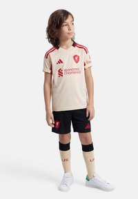 Child standing wearing cream and red Liverpool football kit with black shorts, cream socks, and white sneakers, looking sideways.