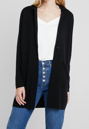 Vrouw die een zwarte lange cardigan met zakken aan de voorkant draagt over een wit V-hals topje en blauwe high-waisted jeans met zichtbare knopen.