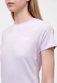 Jeune personne aux longs cheveux blonds portant un t-shirt violet clair à manches courtes avec un petit logo blanc ailé sur la poitrine.