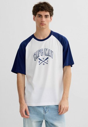 Joven con camiseta de béisbol raglán blanca y azul marino con texto "Cape May" y remos cruzados, combinado con vaqueros azul claro.