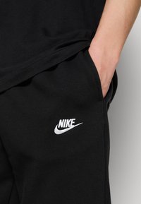 Schwarze Baumwoll-Jogginghosen mit einem weißen, gestickten Nike-Logo auf der linken Tasche. Glatte Textur, lässige Passform und seitliche Taschendetails.