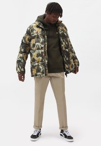 Chaqueta tipo plumón en un patrón de camuflaje verde, beige y marrón; lleva una sudadera verde oscuro debajo; combinada con pantalones beige y zapatillas negras.