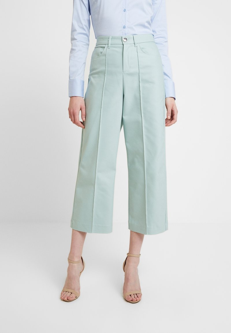Femme portant un pantalon large court taille haute bleu clair et une chemise bleu clair boutonnée, associée à des talons nus à bride à la cheville.