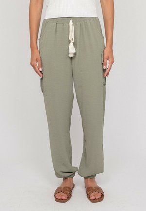 Femme portant un pantalon à cordon de serrage vert olive clair avec poches latérales et des sandales marron à enfiler, debout devant un fond uni.