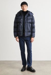 Marinfärgad puffjacka med texturerad yta, avtagbar huva och fickor med dragkedja, kombinerad med blå jeans och svarta skor på en neutral bakgrund.