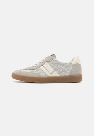 Niski szary i biały sneaker z kwiatową koronkową nakładką i brązową gumową podeszwą, pokazany z boku na białym tle.