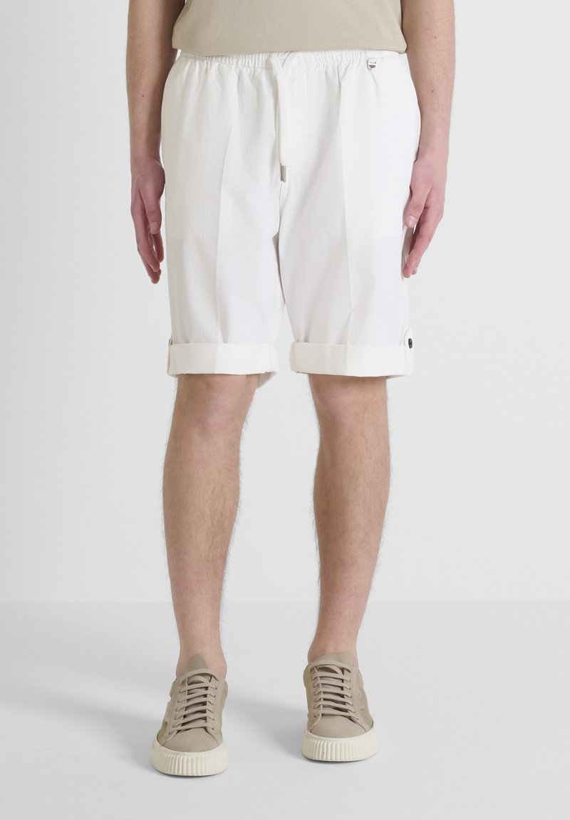 Pantaloni corti bianchi fino al ginocchio con vita regolabile, orlo risvoltato e tasche laterali, realizzati in tessuto leggero. Indossati con sneaker beige.