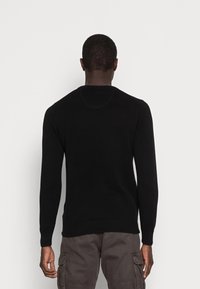 Man draagt een zwart sweater met lange mouwen en donkergrijze cargobroek, staat met zijn rug naar de camera tegen een effen witte achtergrond.