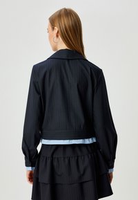Giacca nera a righe con maniche lunghe, colletto a rever e una camicia blu a strati sotto. Texture liscia e vestibilità sartoriale.