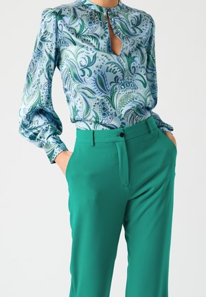 Blusa de manga larga azul claro con patrón de cachemira verde y escote con abertura en forma de ojo de cerradura, combinada con pantalones verdes de tiro alto y sólidos, con las manos en los bolsillos.