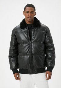 Veste bomber en cuir noir avec fermeture éclair sur le devant, col en fourrure noire, texture matelassée, et bords côtelés aux poignets et à l'ourlet. Chemise blanche en dessous.