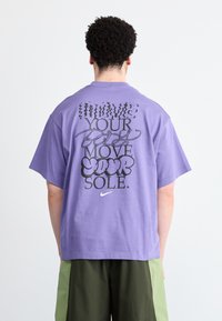 Fialové bavlněné tričko volného střihu. Na zadní straně se nachází černobílý grafický text: "PŘENES SVOU DUŠI." a logo Nike.