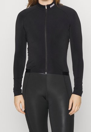 Femme portant une veste de cyclisme noire ajustée à manches longues avec une fermeture éclair frontale et un pantalon de cyclisme noir taille haute.