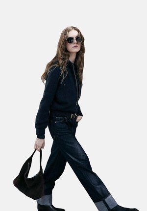 Suéter oscuro, jeans de mezclilla oscuros de talle alto con dobladillos enrollados, bolso negro y gafas de sol redondas. Cabello largo y ondulado con textura natural.