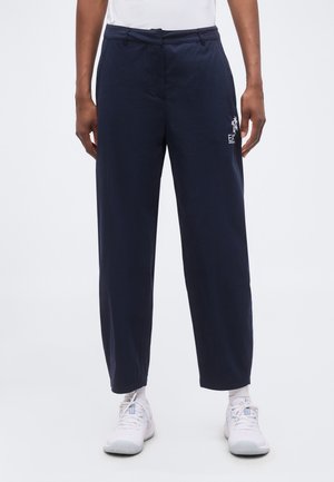 Mężczyzna noszący granatowe proste spodnie z logo z boku, białe skarpetki, białe buty sportowe oraz białą koszulkę wpuszczoną do spodni.