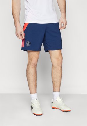 Mężczyzna ubrany w granatowe spodenki Manchester United z czerwonymi paskami, białe skarpetki i białe korki Adidas, stojący na jednolitym tle.