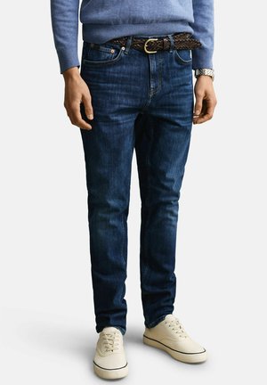 Man draagt donkerblauwe spijkerbroek, gevlochten bruine leren riem, lichtblauwe trui, zilveren horloge en witte casual sneakers, staand tegen een effen achtergrond.