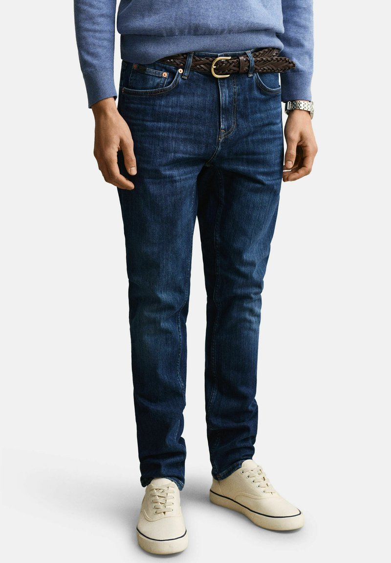 Homme portant un jean bleu foncé, une ceinture en cuir tressé marron, un pull bleu clair, une montre argentée et des baskets blanches décontractées, debout devant un fond uni.
