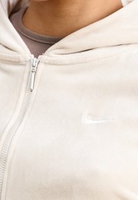 Krémově barevná mikina na zip z měkké tkaniny, s jemným logem Nike na hrudi a přiléhavou kapucí s hladkou texturou.