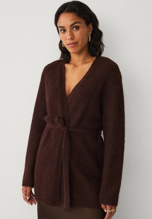Cardigan brun et duveteux avec un design enveloppant et une ceinture à nouer. Les manches longues et un décolleté en V profond créent une silhouette décontractée.