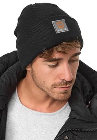 Black knit beanie with a soft texture and a central gray patch featuring the word "JEANSCODE" in orange. Worn with a gray sweater and black jacket.