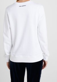 Sudadera blanca de manga larga con dobladillo acanalado. Presenta un logo en negro en la parte posterior del cuello. Textura suave y diseño casual.
