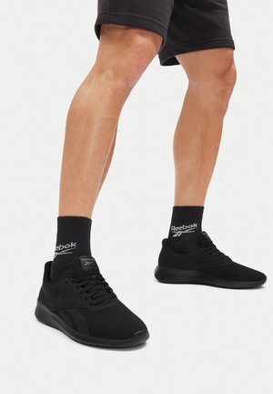 Nohy v černých sportovních botách Reebok, černých ponožkách Reebok a černých šortkách na bílém pozadí.