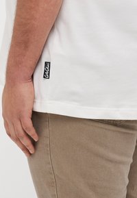 White cotton t-shirt with side seam label. Visible hand resting on khaki fabric, showcasing texture and material contrast.