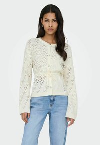 Cardigan en maille crème avec un motif texturé, des boutons sur le devant et une ceinture nouée à la taille. Manches longues et coupe décontractée mettent en valeur son design.