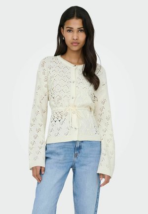 Cardigan en maille crème avec un motif texturé, des boutons sur le devant et une ceinture nouée à la taille. Manches longues et coupe décontractée mettent en valeur son design.