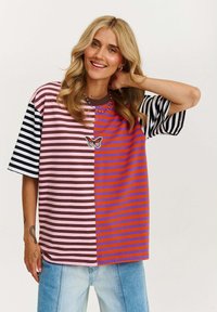 Pasiasty, oversize t-shirt z różowymi, brązowymi, fioletowymi i pomarańczowymi sekcjami, z motywem motyla i okrągłym dekoltem. Materiał bawełniany.